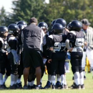 A man in a black windbreaker stands in the huddle of a youth football team with black uniforms and helmets.