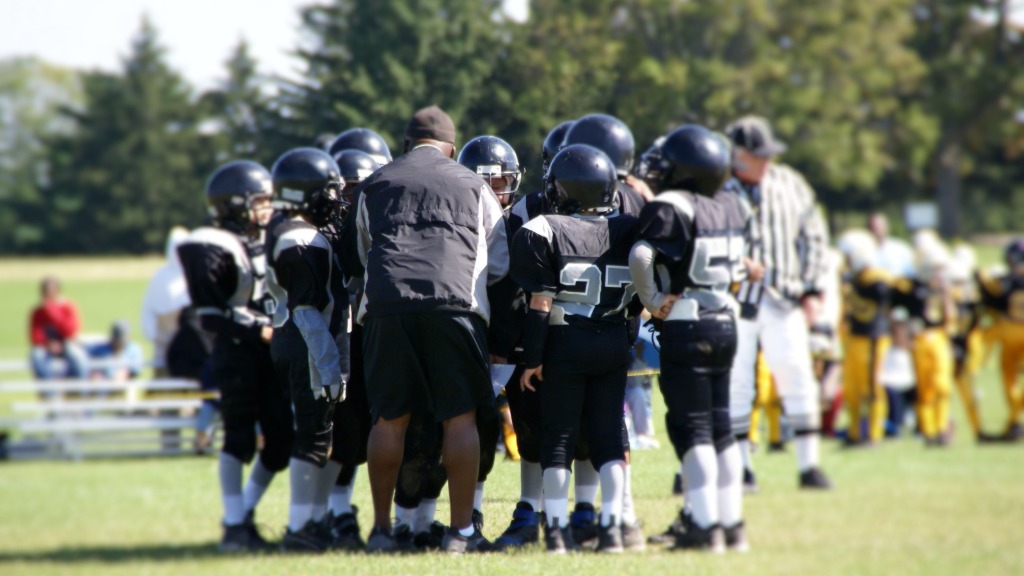 A man in a black windbreaker stands in the huddle of a youth football team with black uniforms and helmets.