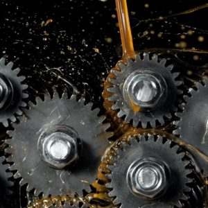 Multiple tooth gear wheels are spinning. Gear oil is spilled onto the gears and splattered against a black background.