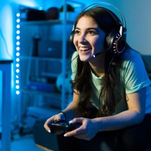 A young woman sits on the edge of a couch in a dimly lit room, wearing headphones and holding a gaming controller.