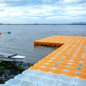 A blue and orange plastic floating dock on a shore with a canoe next to it and shoreline in the background.