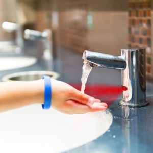 A close-up view shows a person wearing a blue bracelet placing their hand under the automatic faucet in a restroom.