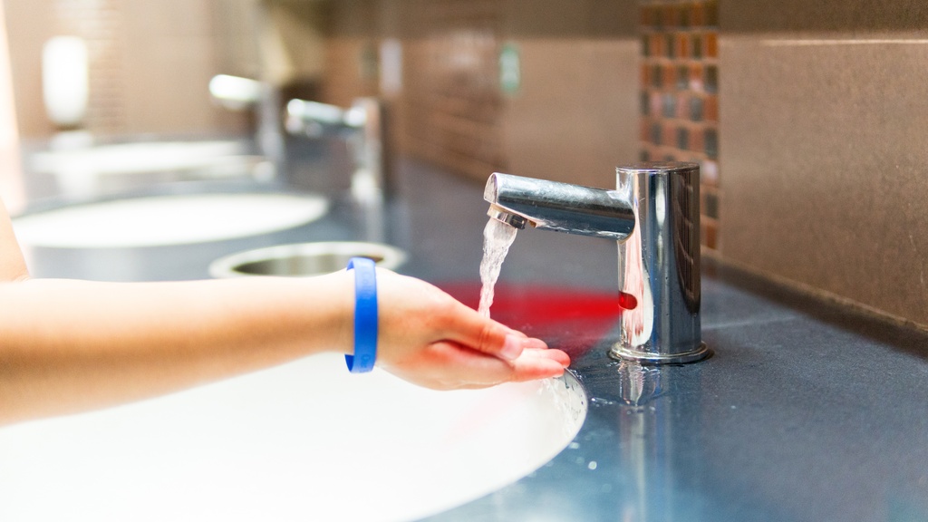 A close-up view shows a person wearing a blue bracelet placing their hand under the automatic faucet in a restroom.