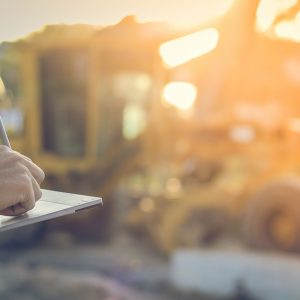 A close-up of a worker writing on an electronic tablet with construction equipment blurred in the background.