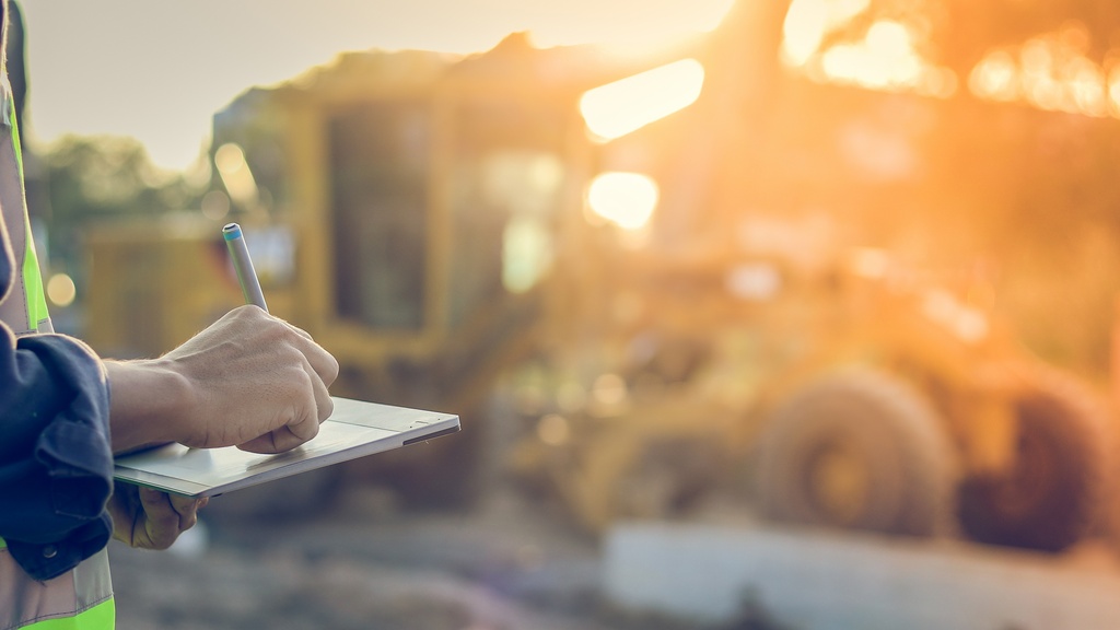 A close-up of a worker writing on an electronic tablet with construction equipment blurred in the background.
