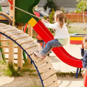 Children playing at a playground together with an adult behind them. One girl is climbing up a ramp with a rope.