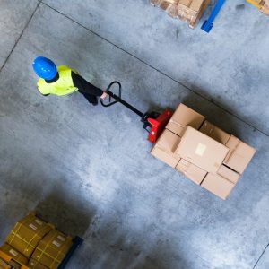 A person in a blue hardhat is pulling a pallet jack loaded with boxes through a warehouse.