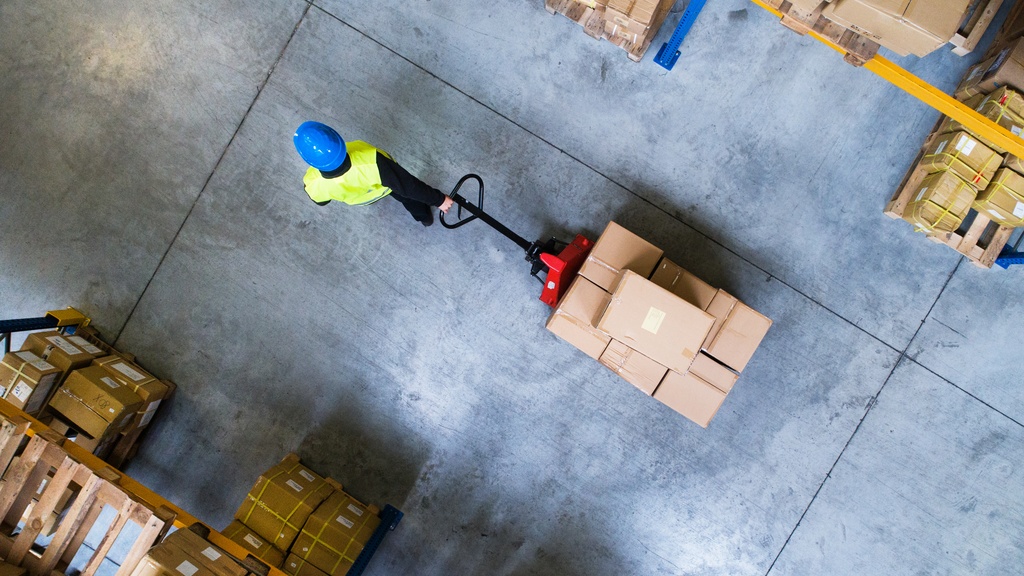 A person in a blue hardhat is pulling a pallet jack loaded with boxes through a warehouse.
