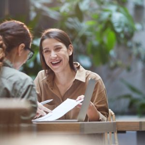 Two friends spend time together at an outdoor cafe. One of the friends is laughing as they look at a document.