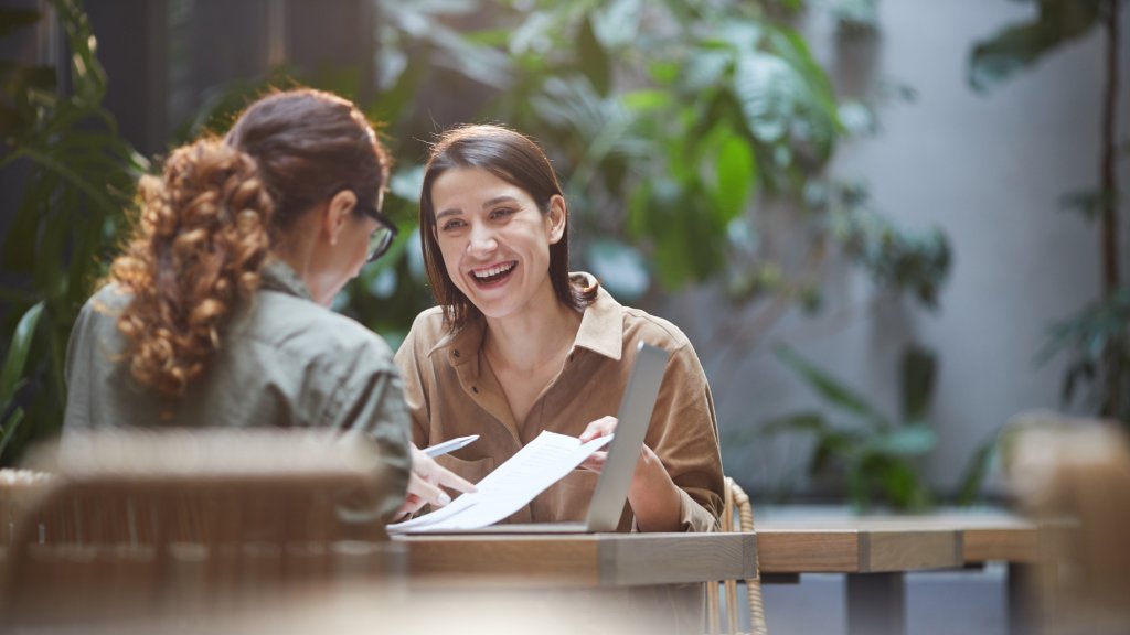 Two friends spend time together at an outdoor cafe. One of the friends is laughing as they look at a document.