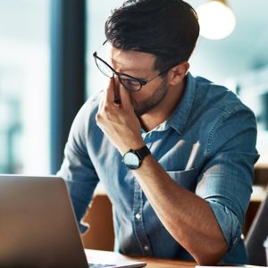 A man sits at his desk at work and rubs the bridge of his nose in frustration. He sits in front of a laptop.