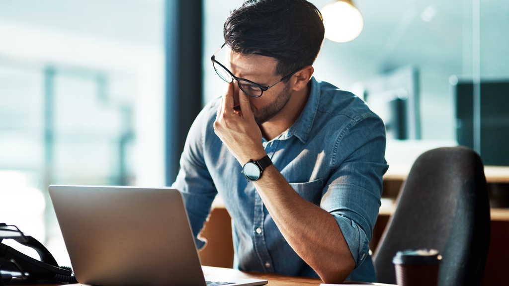 A man sits at his desk at work and rubs the bridge of his nose in frustration. He sits in front of a laptop.