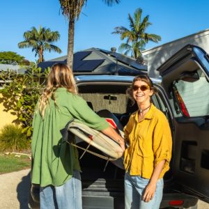 Two women are standing behind a hatch-back car with he hatch open. One woman is looking at the camera.