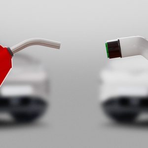 A red gasoline nozzle and electric vehicle charging plug facing each other with blurred cars in the background.