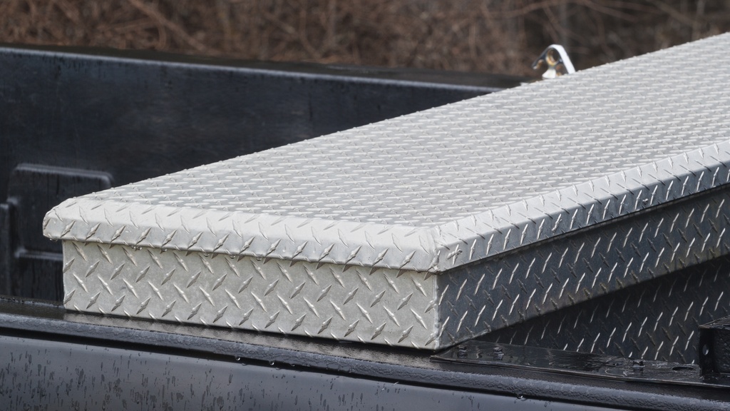 A close-up of a large, silver toolbox resting across the back of a truck bed. The truck is black.