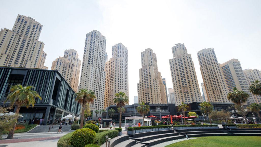 A green public park with palm trees, benches, and a paved walkway all sitting in front of a bunch of tall buildings.