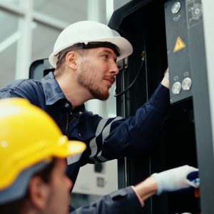 Two engineers wearing gloves and hard hats working on the electrical system inside a brightly lit industrial workplace.