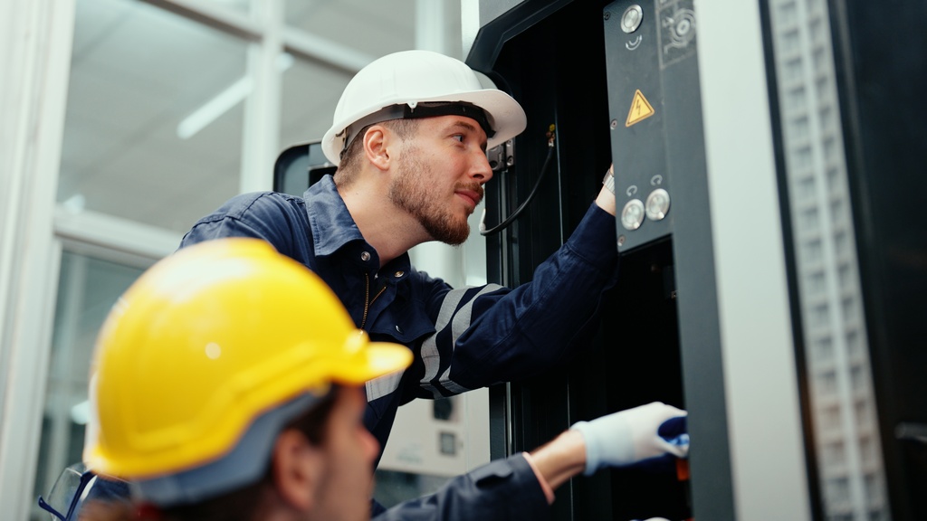 Two engineers wearing gloves and hard hats working on the electrical system inside a brightly lit industrial workplace.