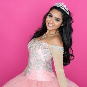 A smiling young woman wearing a jeweled crown and pink formal dance dress against a bright pink background.