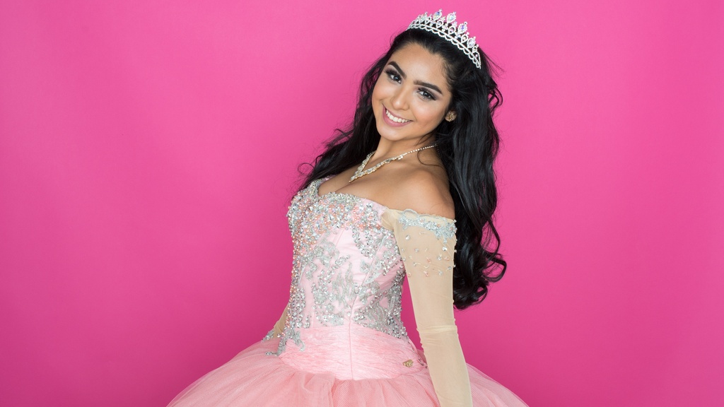 A smiling young woman wearing a jeweled crown and pink formal dance dress against a bright pink background.
