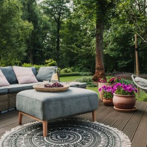An outdoor space with a wooden deck, a round area rug, a swinging chair, and a large couch next to a forest.