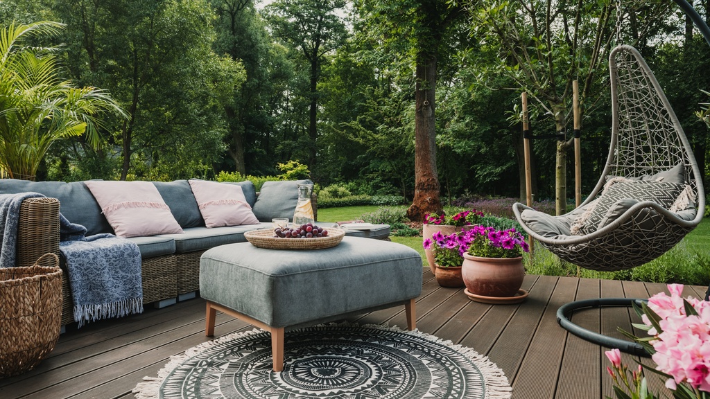 An outdoor space with a wooden deck, a round area rug, a swinging chair, and a large couch next to a forest.
