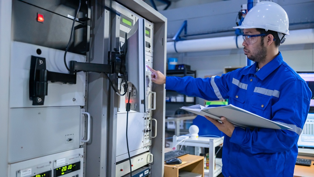 An industrial worker in a blue jumpsuit and white hard hat interacts with machinery while holding a document binder.