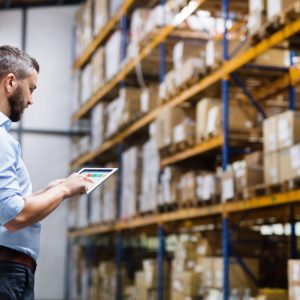 A man wearing a button-down shirt and dark pants uses a tablet in a warehouse aisle beside tall racks and stacked boxes.