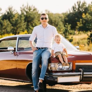 Adult wearing sunglasses and a child sit on the front of a vintage brown car parked near trees in warm light.