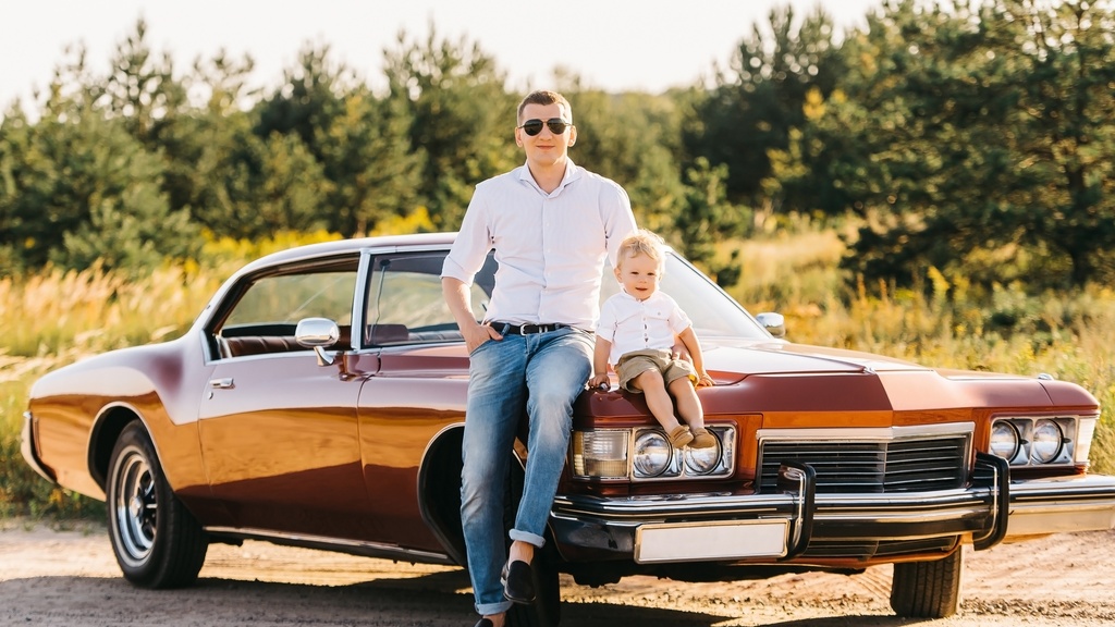 Adult wearing sunglasses and a child sit on the front of a vintage brown car parked near trees in warm light.