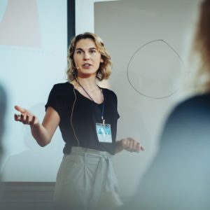 A woman wearing a lanyard speaking in front of a projector screen and whiteboard, with blurred people in the foreground.