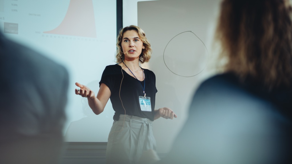 A woman wearing a lanyard speaking in front of a projector screen and whiteboard, with blurred people in the foreground.