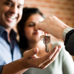 A smiling couple standing next to each other indoors while one of them holds out their hand for a set of keys.