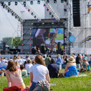 Crowd of festival attendees sitting on grass watching a live outdoor music concert with a large lit stage and big screen.