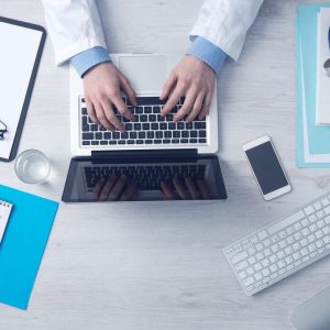 overhead shot of medical professional typing on laptop keyboard with paper and clipboard on the table