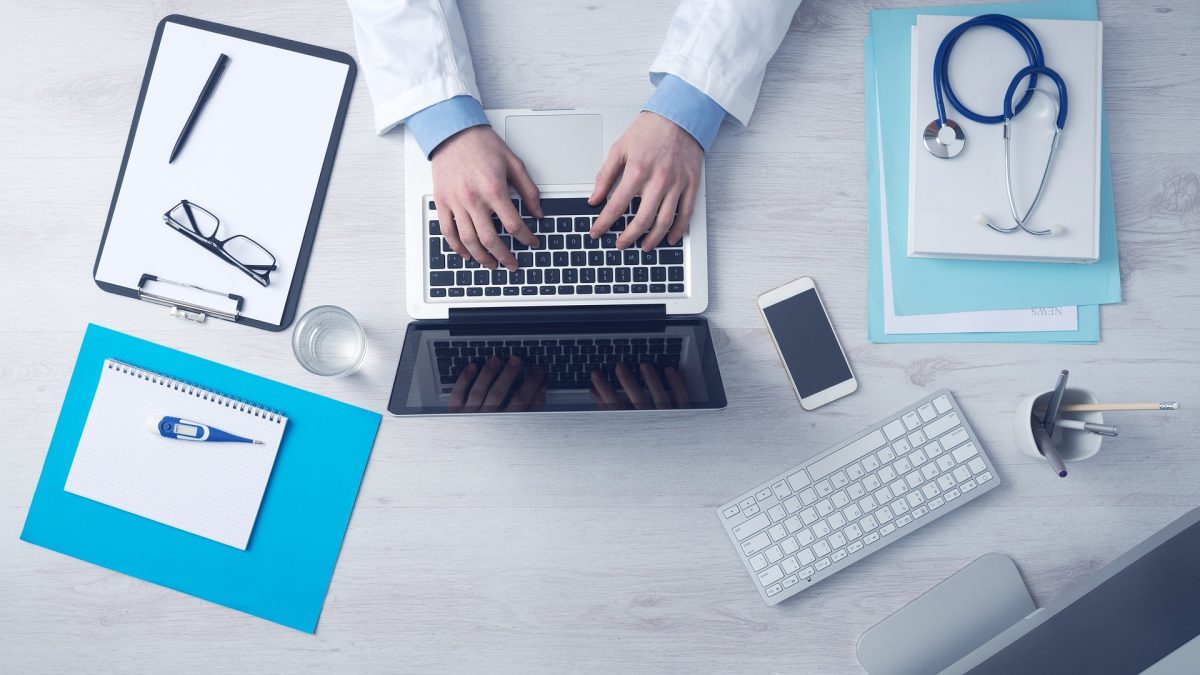 overhead shot of medical professional typing on laptop keyboard with paper and clipboard on the table