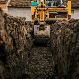 A backhoe bucket digging into brown soil beside a trench, with loose dirt and construction ground in view.