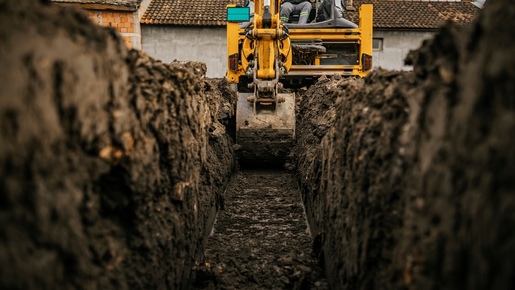 A backhoe bucket digging into brown soil beside a trench, with loose dirt and construction ground in view.