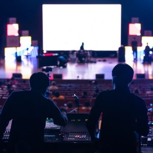 Two silhouetted people working at audio control boards while facing a lit stage and rows of seats in a dark venue.