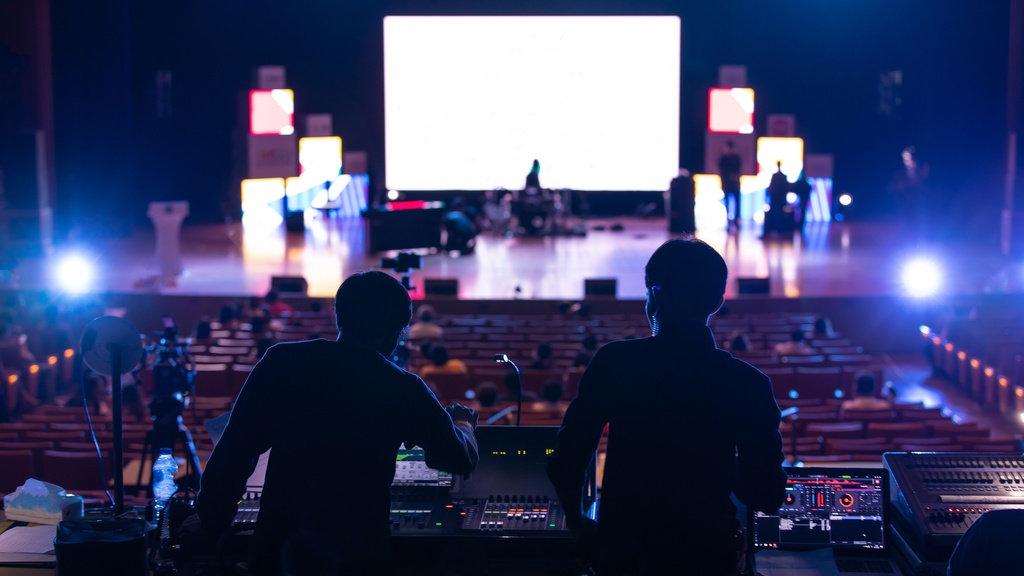 Two silhouetted people working at audio control boards while facing a lit stage and rows of seats in a dark venue.