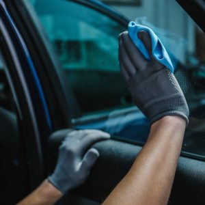 Mechanic wearing gloves using a blue rag to wipe down dark tinted windows on a car parked outside during the daytime.
