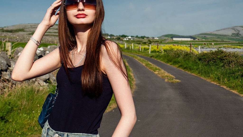 A young woman in wide leg jeans and a black tank top adjusts her sunglasses on a sunny rural road with green hills behind her.