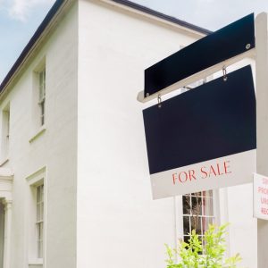 A for-sale sign hangs in front of a tall, white mansion. It has two stories, multiple windows, and bushes.
