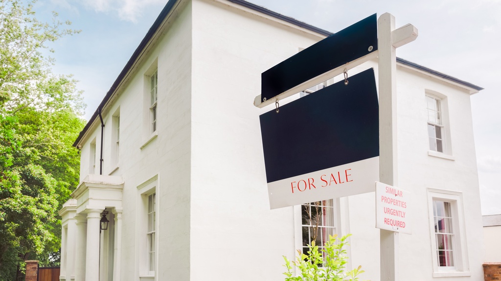 A for-sale sign hangs in front of a tall, white mansion. It has two stories, multiple windows, and bushes.