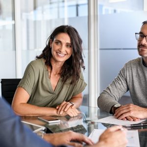 Two people sit side by side at a conference table while another person faces them. Documents are on the table.