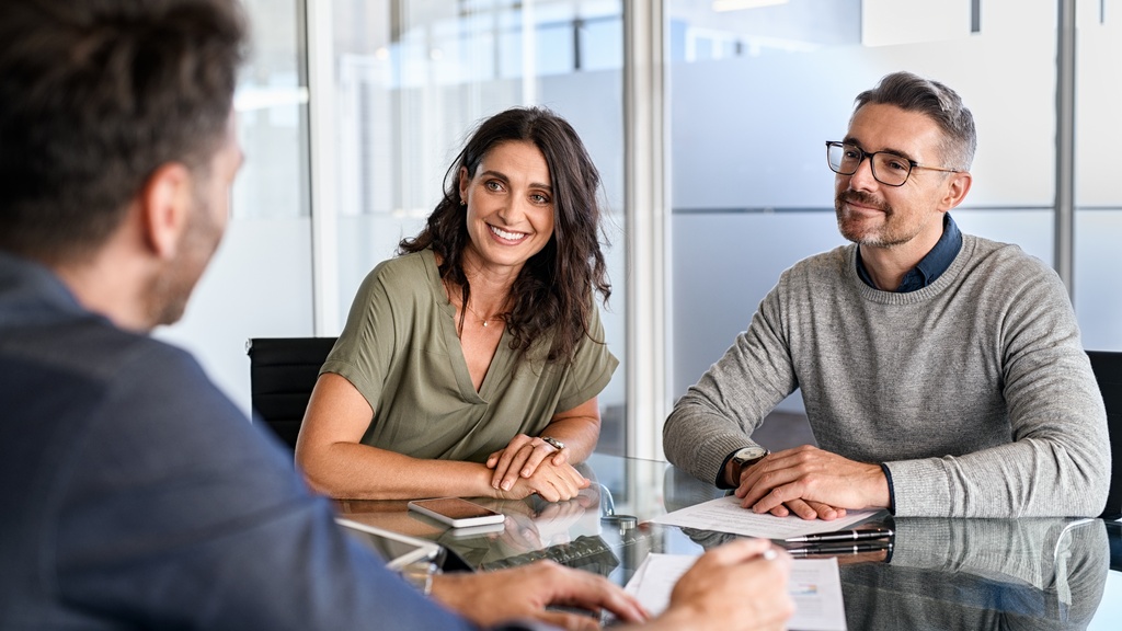 Two people sit side by side at a conference table while another person faces them. Documents are on the table.