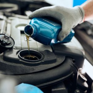 A close-up of a hand with a glove holding a blue bottle of motor oil and pouring it into a car's engine reservoir.