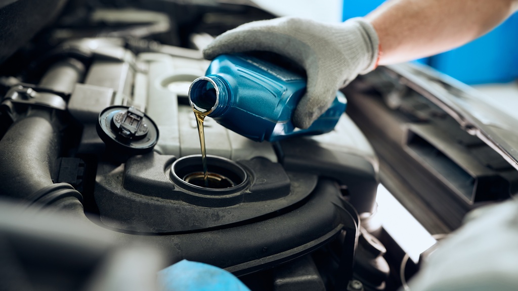 A close-up of a hand with a glove holding a blue bottle of motor oil and pouring it into a car's engine reservoir.