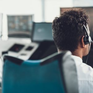 Two men wearing headsets sitting on ergonomic chairs behind a desk with multiple monitors and a large window.