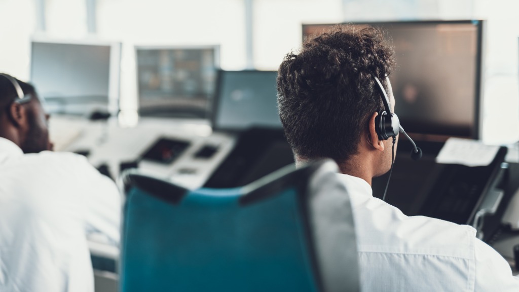 Two men wearing headsets sitting on ergonomic chairs behind a desk with multiple monitors and a large window.